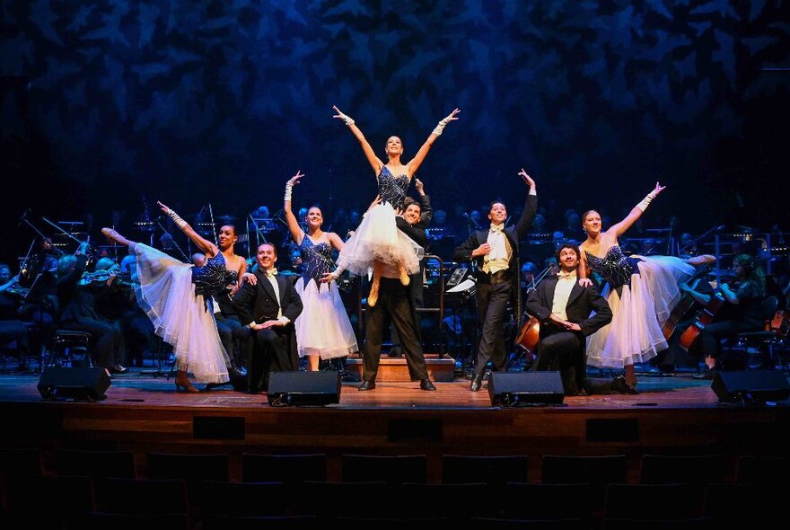 Dancers performing on stage in a group of men wearing tuxedos and women wearing tulle gowns.