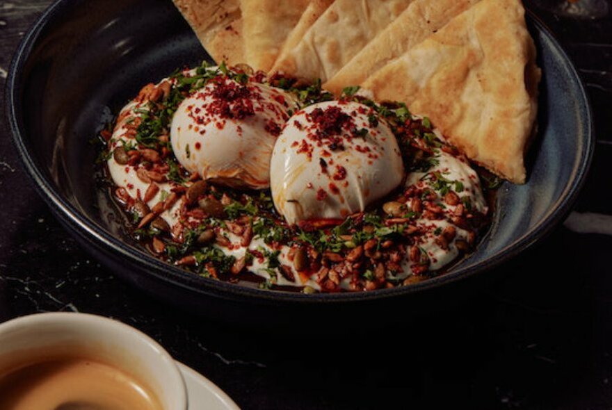 A cup of coffee next to a dish of poached eggs with bread.