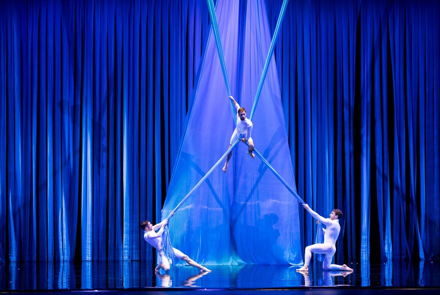 A circus performer balancing between two lengths of fabric, each being held at the bottom by another dancer.
