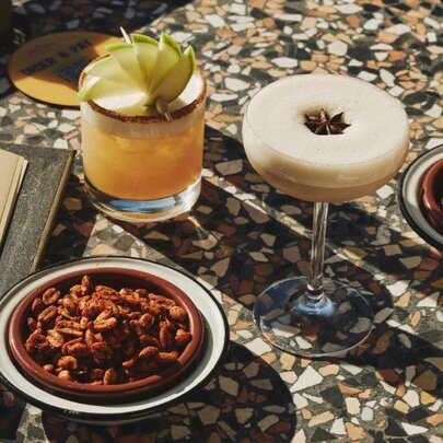 Two cocktails on a table top with a bowl of nibbles nearby and a menu.