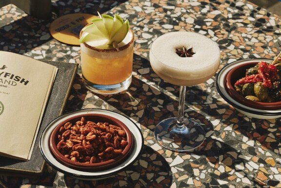 Two cocktails on a table top with a bowl of nibbles nearby and a menu.