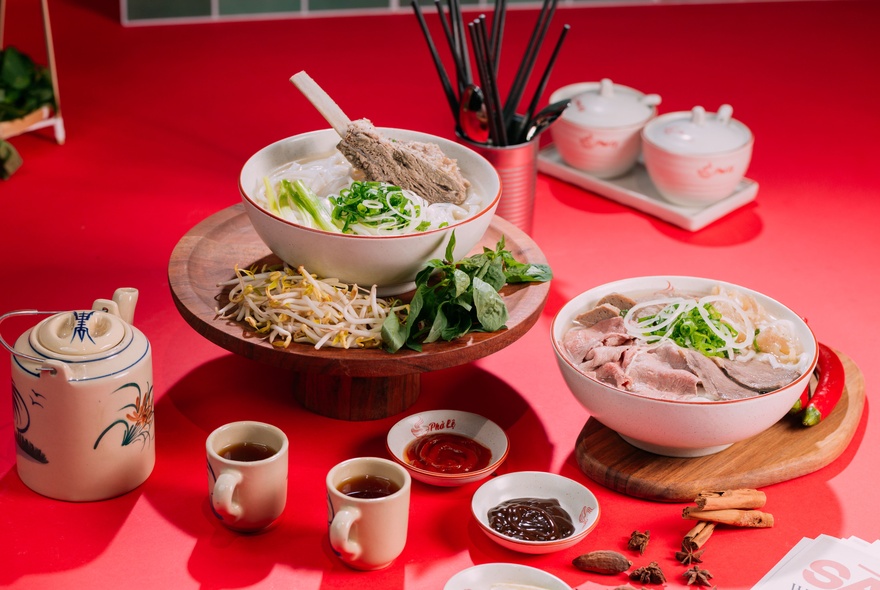 Bowls of pho, a teapot and cups on a red hued table setting with condiments. 