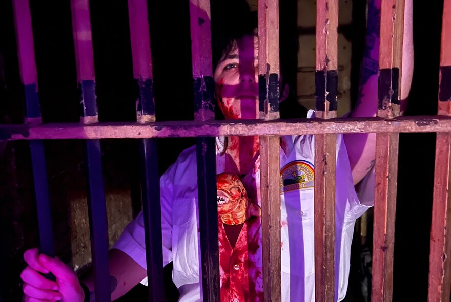An individual behind dark vertical prison bars under purple and reddish dramatic lighting, wearing a light-coloured shirt with an emblem on the chest.