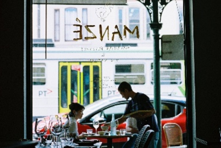 Restaurant window looking out onto a tram passing by, person seated at outdoor table being served wine and food.