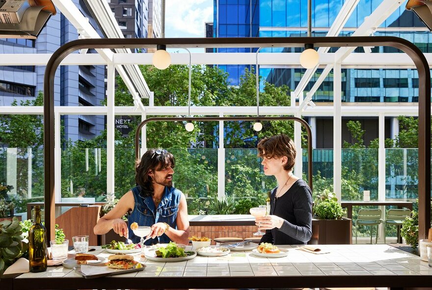 Two friends enjoying share plates and cocktails on a rooftop bar with industrial lighting installations.