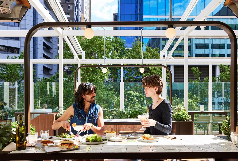 Two friends enjoying share plates and cocktails on a rooftop bar with industrial lighting installations.