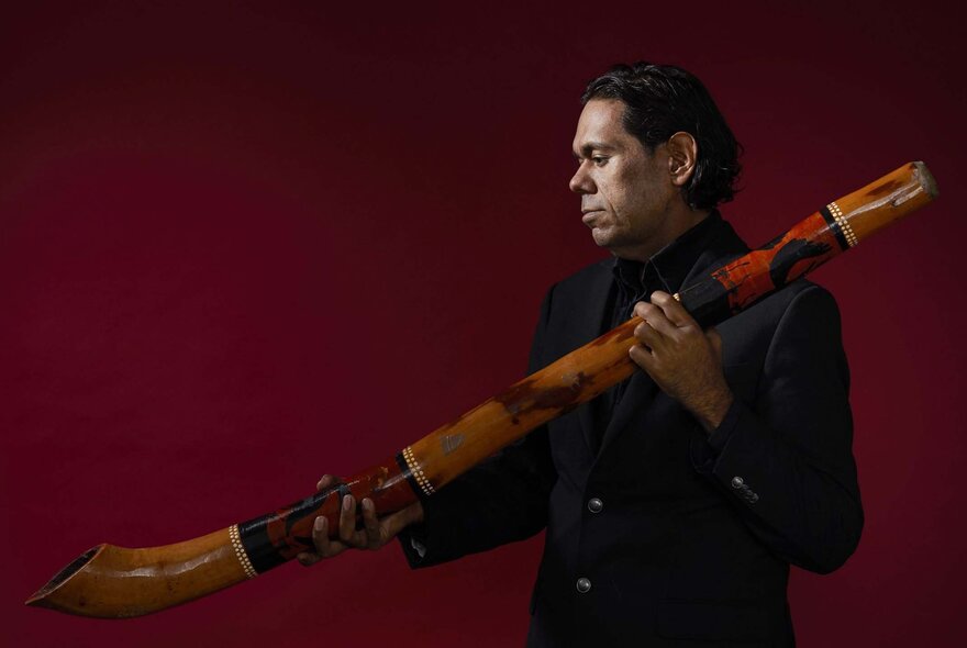Master yidaki artist William Barton holding his instrument and staring sideways against a deep red background. 