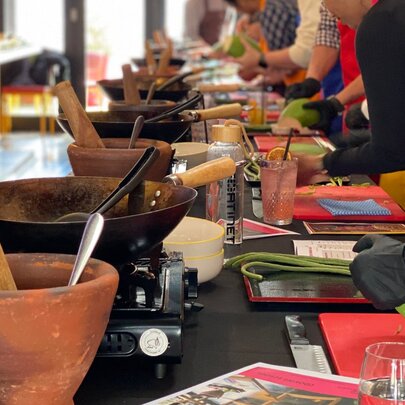A black kitchen workbench with mortars and pestles lined up with woks, people leaning over the nech preparing a Thai vegetable for cooking.