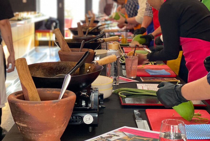 A black kitchen workbench with mortars and pestles lined up with woks, people leaning over the nech preparing a Thai vegetable for cooking.