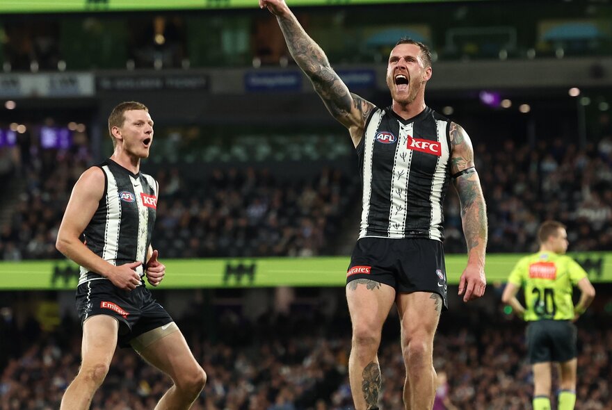 Two AFL Collingwood players on a sports ground cheering with the fans in the stadium around them.