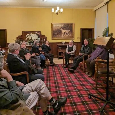 A seated audience watching a panel discussion with a moderator standing at a lecturn, in a room with red tartan carpet and yellow walls.