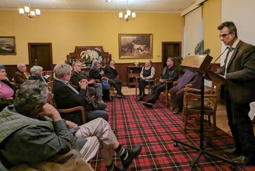 A seated audience watching a panel discussion with a moderator standing at a lecturn, in a room with red tartan carpet and yellow walls.