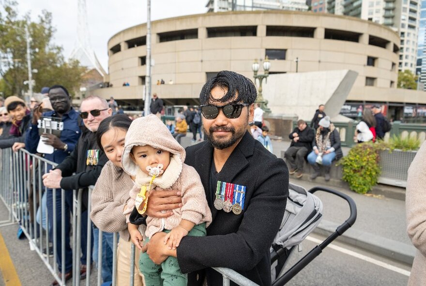 A man wearing medals holding a baby along the railing at the ANZAC Day march in Melbourne.