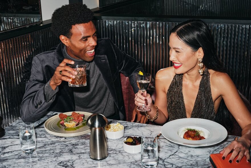 A glamorous couple enjoying a drink and gourmet food at a shiny booth with a marble table.