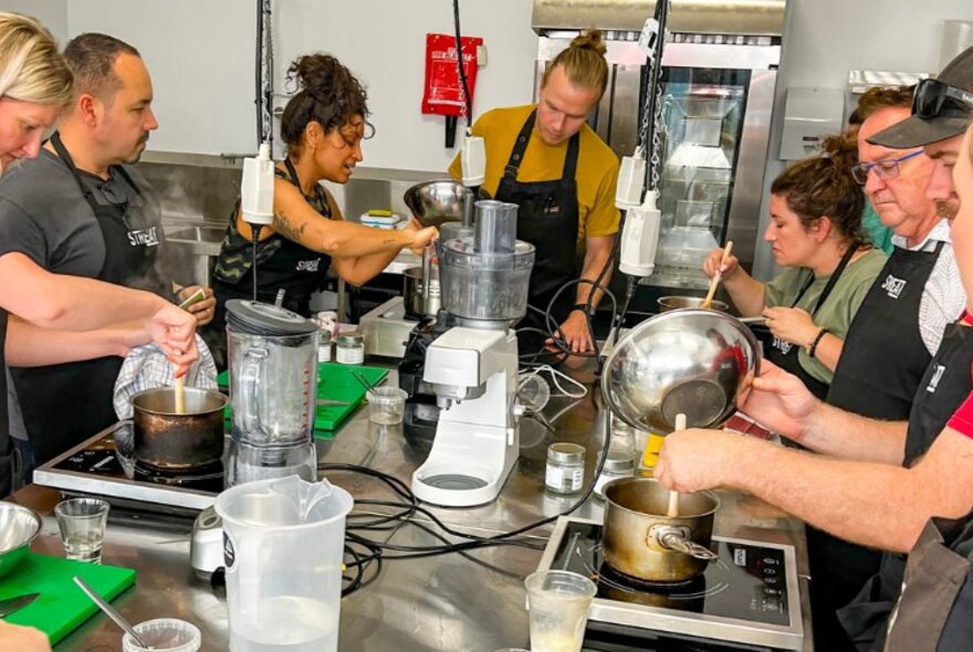A group of people participating in a cooking class, standing around a table in front of individual electric hotplates and stirring saucepans.