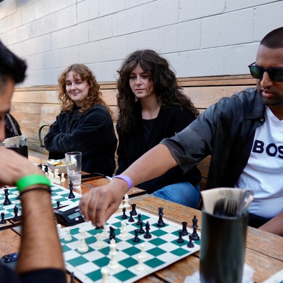 Chess players playing a game at an outdoor table while two women look on.
