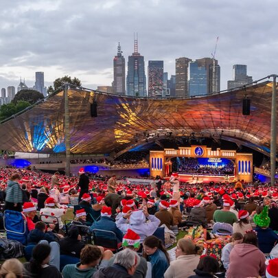 A massive outdoor crowd, many wearing red festive Santa hats gathers for an evening Carols by Candlelight concert at the Sidney Myer Music Bowl with the city skyline visible behind the illuminated canopy stage.