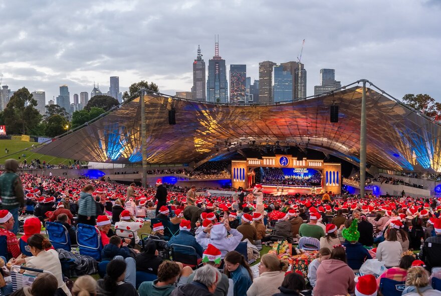A massive outdoor crowd, many wearing red festive Santa hats gathers for an evening Carols by Candlelight concert at the Sidney Myer Music Bowl with the city skyline visible behind the illuminated canopy stage.