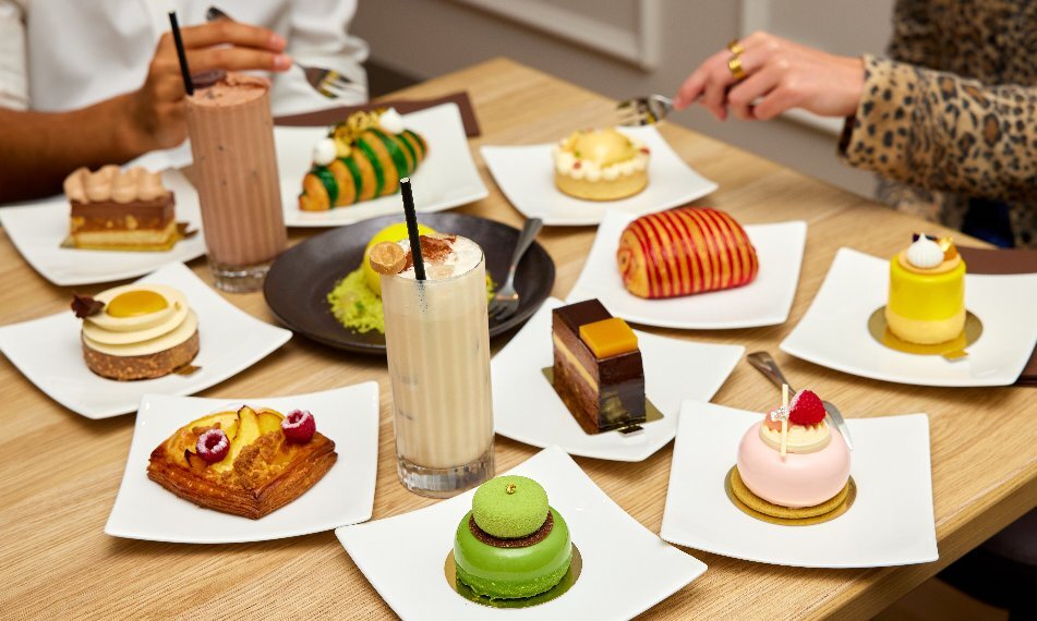 Two people enjoying a wide assortment of cakes, pastries and sweets on white square plates with two iced milk drinks.