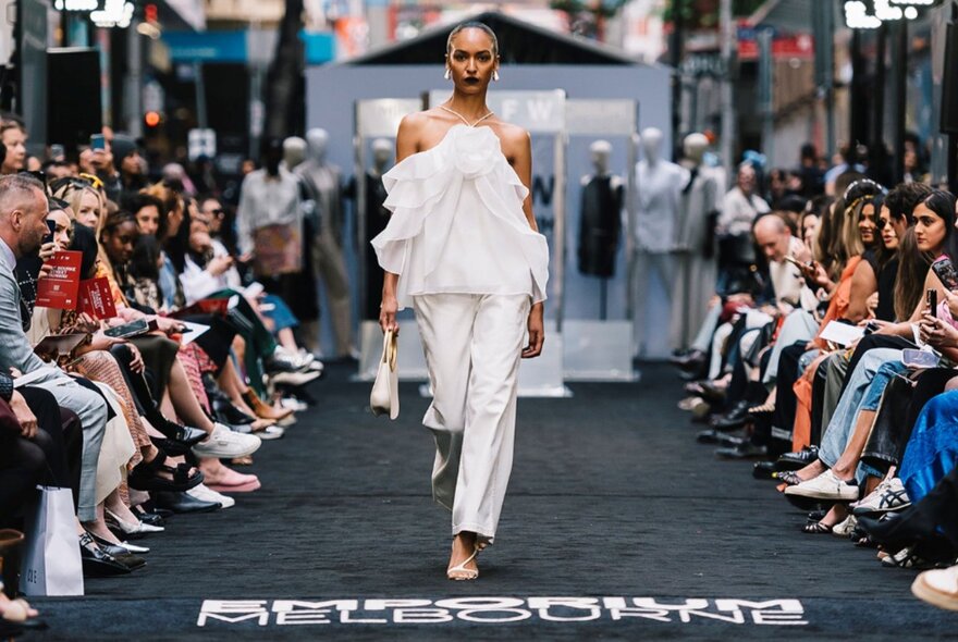 Runway model wearing white trouser suit walking past seated onlookers.