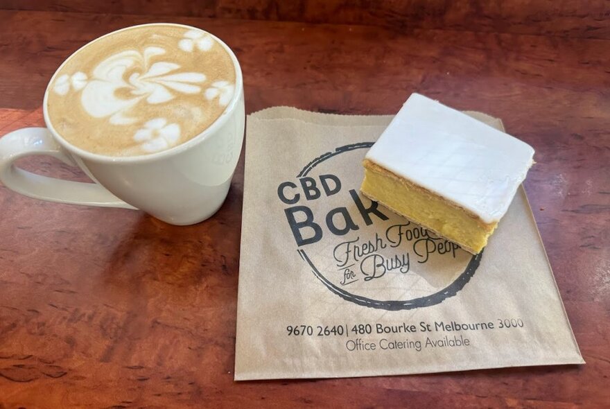 A coffee in a white mug next to a paper bag with a bakery logo printed on it and a vanilla slice on top.