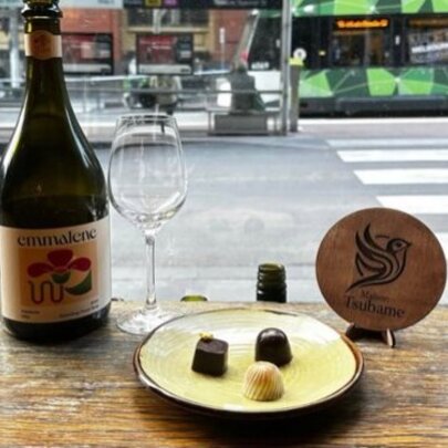 A bottle of wine next to an empty wine glass on a wooden table, and a small plate with three pieces of chocolate on it; a tram and street scene is visible through the window.