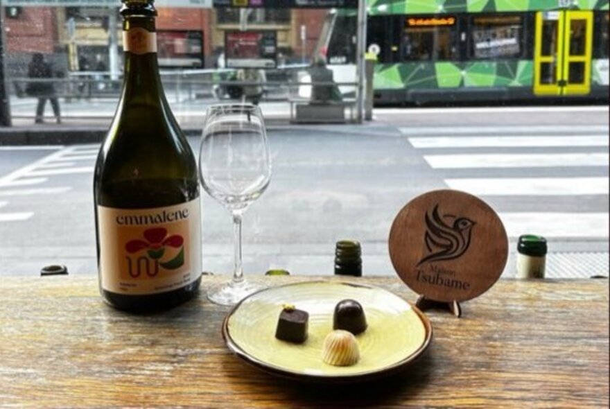 A bottle of wine next to an empty wine glass on a wooden table, and a small plate with three pieces of chocolate on it; a tram and street scene is visible through the window.