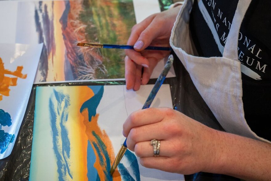 A person wearing an apron, seated at a table as part of am art workshop, using a paintbrush to apply paint to paper.