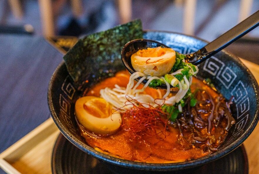 Black bowl of ramen on a tray topped with an onsen egg, wood ear mushrooms, chilli threads and spring onion.