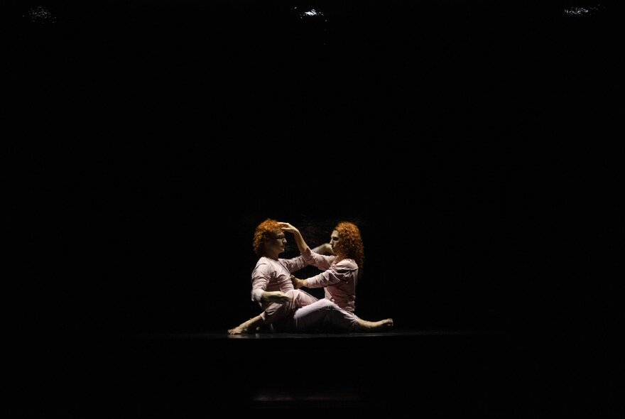 A scene from the contemporary dance work Red by Australian company Dancenorth, with two dancers sitting on a dark stage with legs interlocked.
