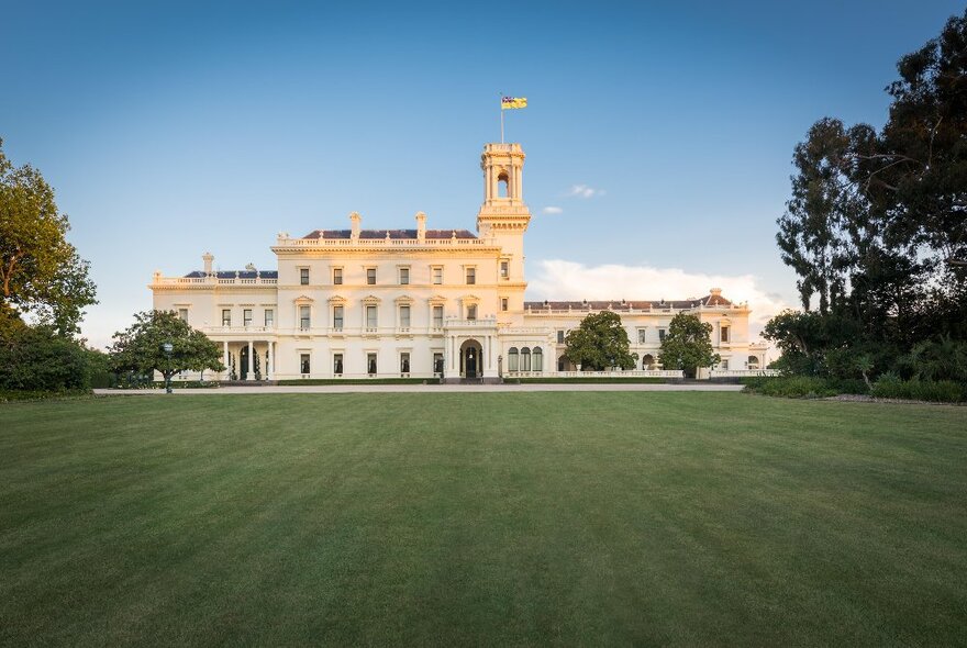 Government House with flag flying from turret, long stretch of lawn and trees in the foreground.