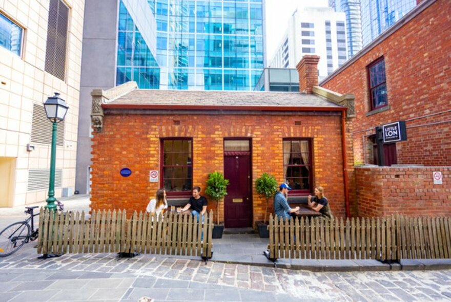 Two couples drinking outside an old red brick cottage with a picket fence in the city. 