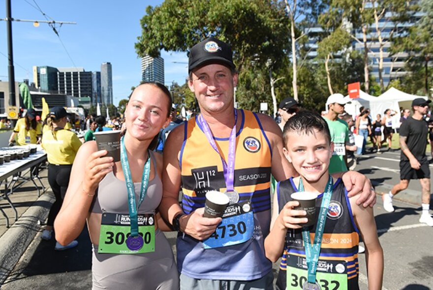 Three run participants standing in a group together outdoors, post run, each wearing completion medals, holding paper cups and smiling at the camera.