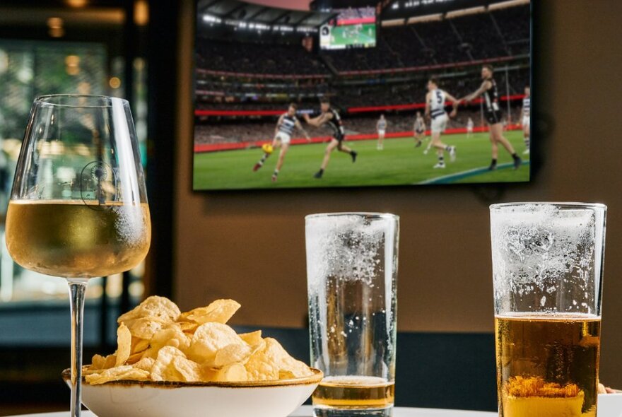 Large screen showing an AFL football game, with glasses of beer and wine and a bowl of crisps in the foreground.