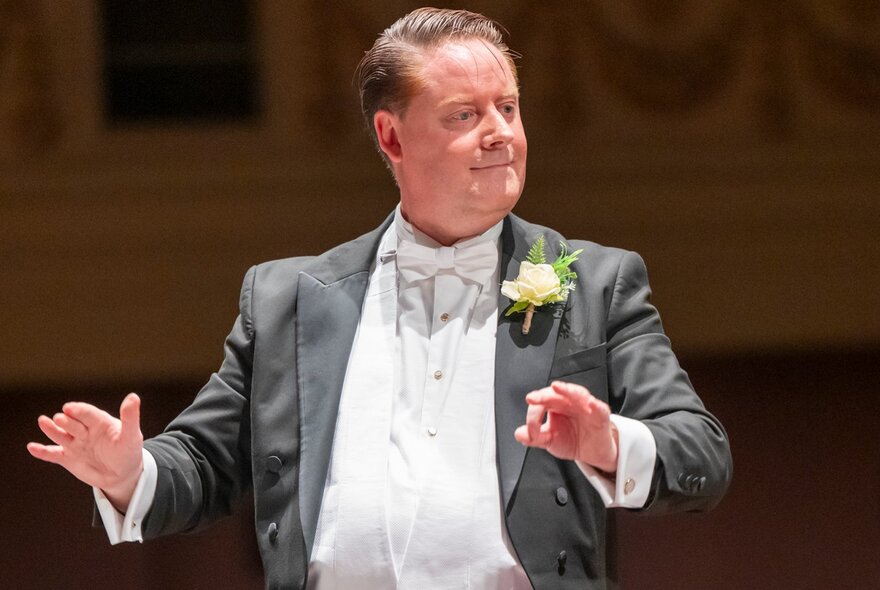 A middle-aged man with short brown hair, performing as a conductor, wearing a formal black tuxedo jacket over a white dress shirt and a white bow tie.