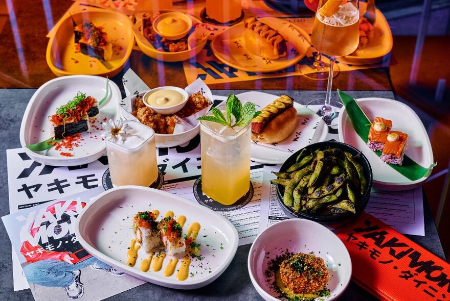 Several plates of Japanese food arranged on a restaurant dining table, with a few cocktails and menus.