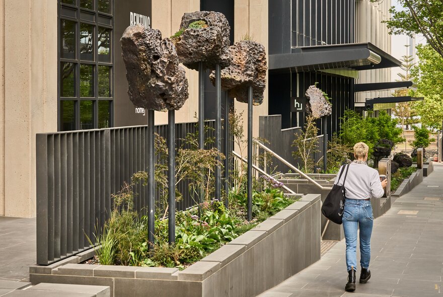 Three bronze artworks on poles in a garden bed in front of a city building, with a pedestrian walking past.