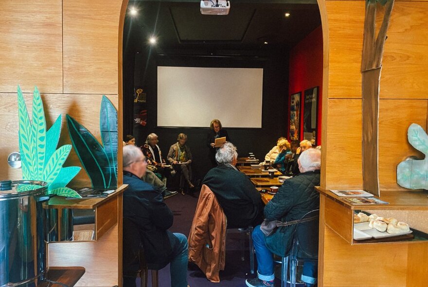 People sitting around a table in a room at Cinema Nova, listening to a person hosting a talk, a large screen on the rear wall.