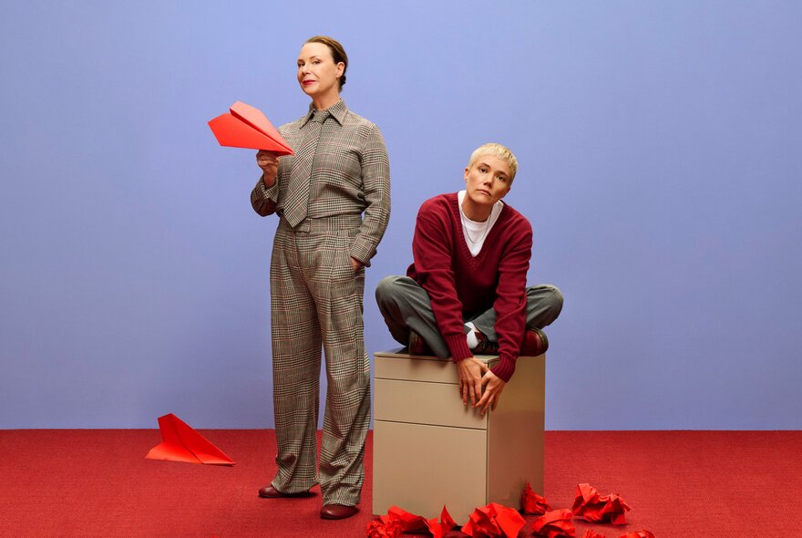 Two actors posing – one standing with a paper plane and the other sitting cross-legged on a box – on a room with a red floor and blue wall. 