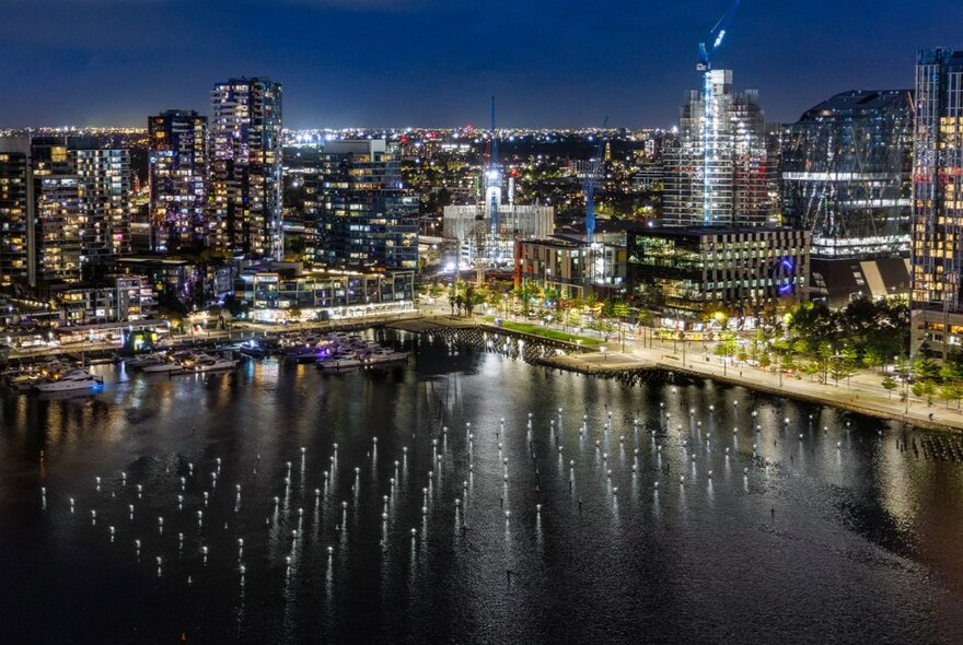 Night aerial view of the Melbourne Docklands skyline with the calm waters of Victoria Harbour and a public art installation of illuminated buoys in rows in the foreground. 