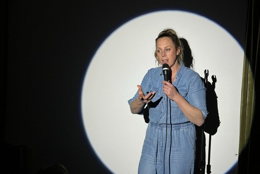 A stand up comedian on a stage, holding a microphone, performing in a spotlight.