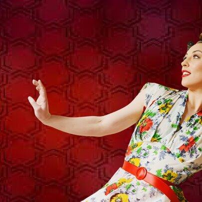Woman wearing a floral-patterned retro dress with red belt, her arm outstretched.