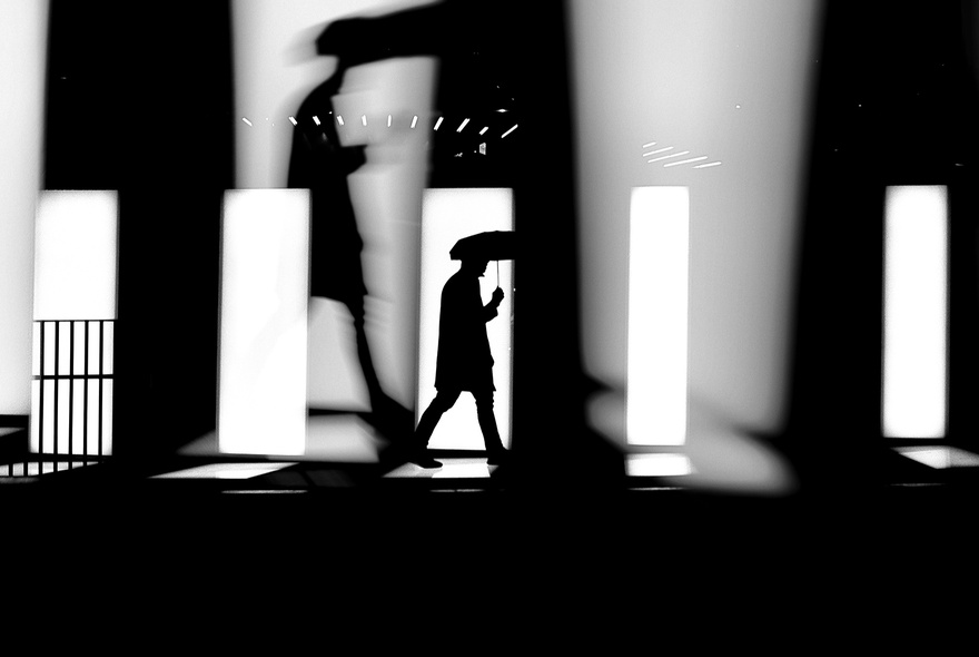 A black-and-white street photograph of a high-contrast scene focused on silhouettes and shadows. 
