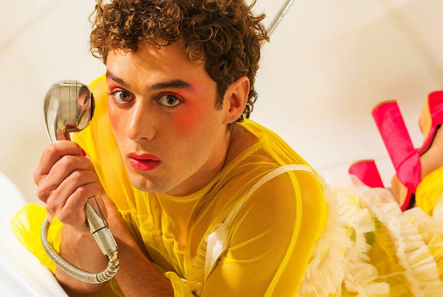 A radiantly-beautiful gender-fluid person wearing a yellow see-through top, pink makeup and high heels, holding a gold shower head like a corded phone.