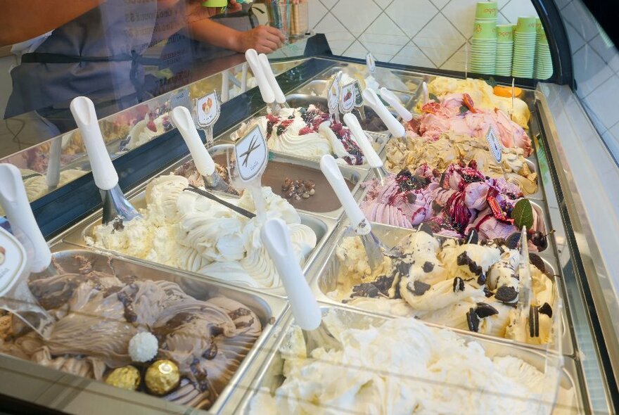 Tubs of different gelato in a refrigerated glass display cabinet.