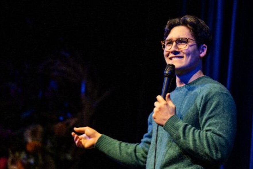 A comedian talking into a microphone they hold in their hand, while standing on a stage in front of an audience.