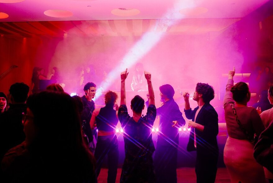 People dancing while in the hazy background a band appears to be performing on a stage, with pink filtered lights illuminating the stage and surrounds.