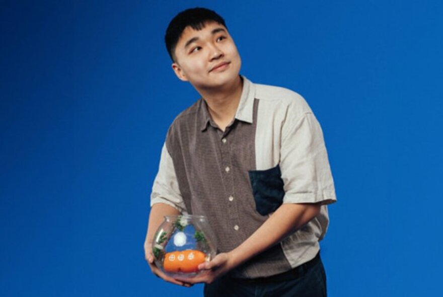 Comedian Jin Hao Li, a young man looking upwards and smiling, short dark hair, wearing a casual short sleeve shirt and holding a glass bowl with an orange object in it, posed against a blue backdrop.