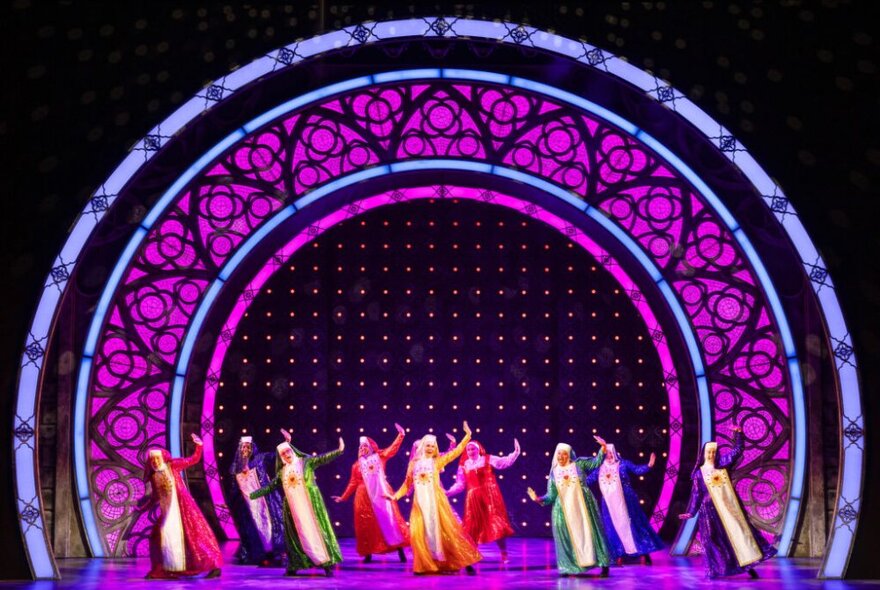 A group of nuns in colourful habits, singing on stage with a large purple stained glass set decoration behind them.