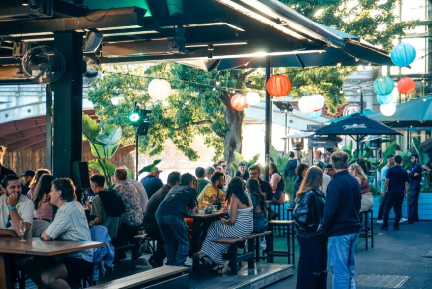 People seated at tables on the wharf outside Riverland Bar, the area set up with large umbrellas and festive lanterns. 